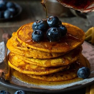 A stack of pumpkin pancakes topped with blueberries with syrup being poured