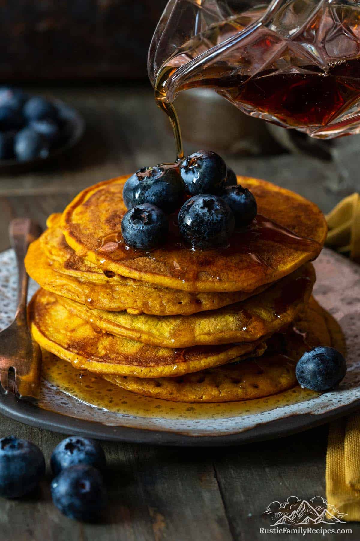 A stack of pumpkin pancakes topped with blueberries with syrup being poured