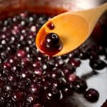 A wooden spoon with blueberry sauce in front of a skillet with cooking sauce