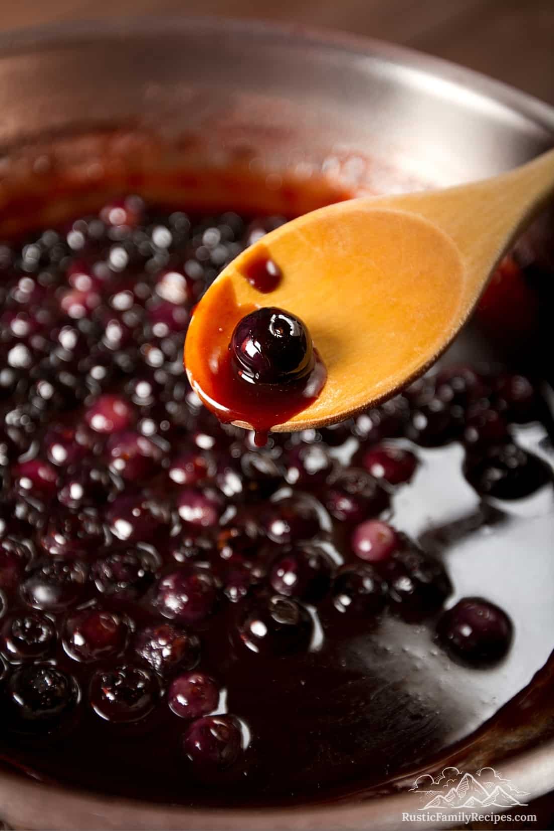 A wooden spoon with blueberry sauce in front of a skillet with cooking sauce