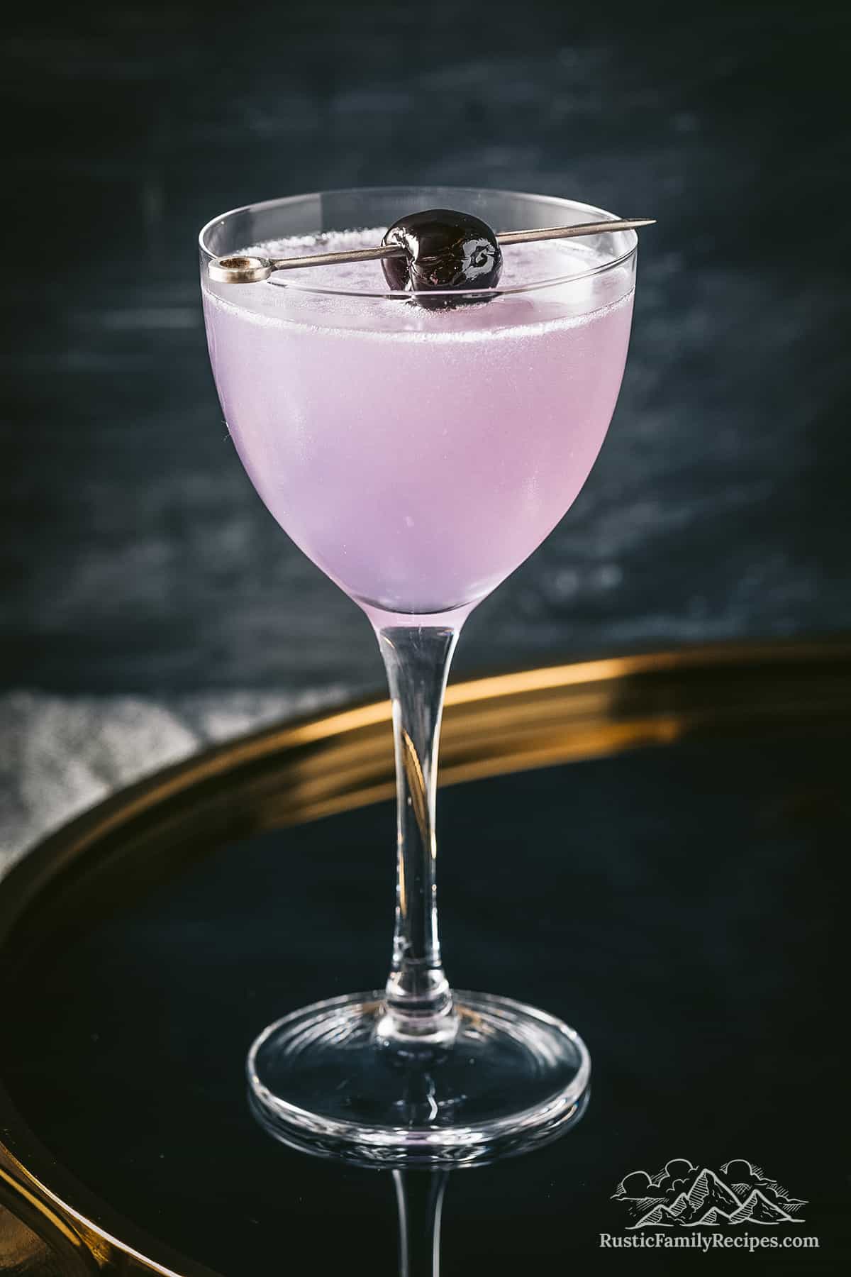A cocktail glass filled with a purple drink and topped with a brandied cherry