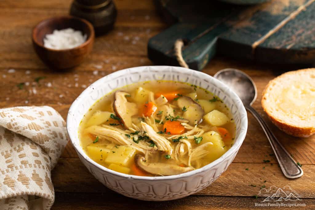 A bowl of ip chicken soup on a wood table next to a spoon and slice of bread.