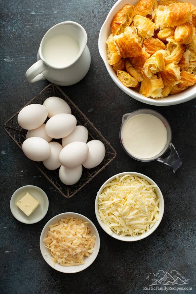 Bowls of cheese next to a basket of eggs, a bowl with croissant pieces, and a measuring cup with cream
