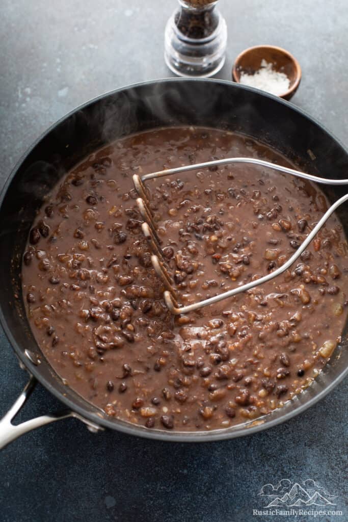 A potato masher mashing cooked black beans in a pan