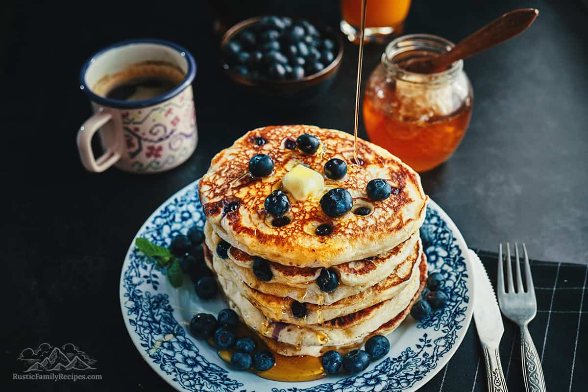 Pouring honey on a stack of multigrain pancakes with butter and blueberries.