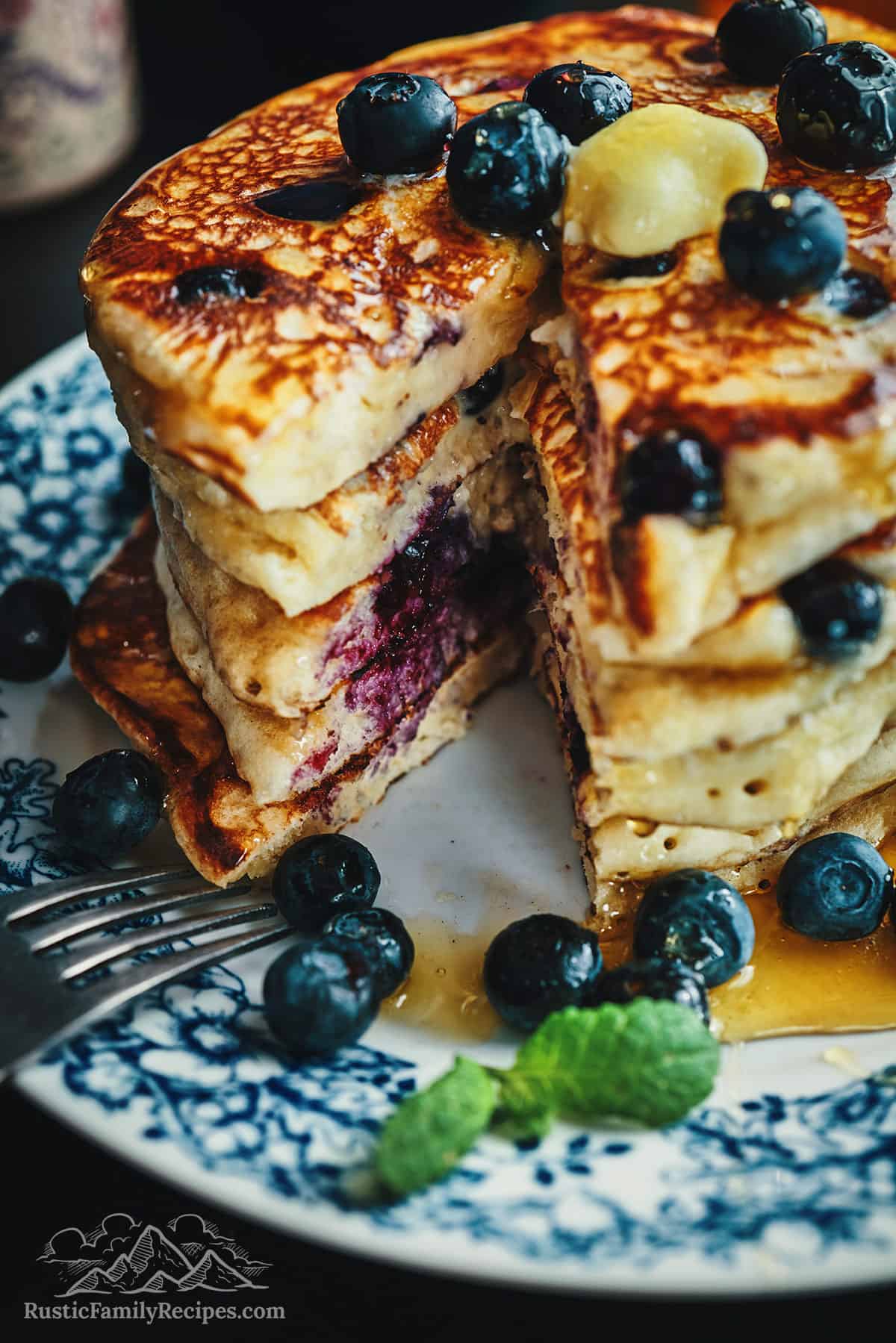 A tall stack of multigrain pancakes with a wedge cut out, topped with blueberries and butter.