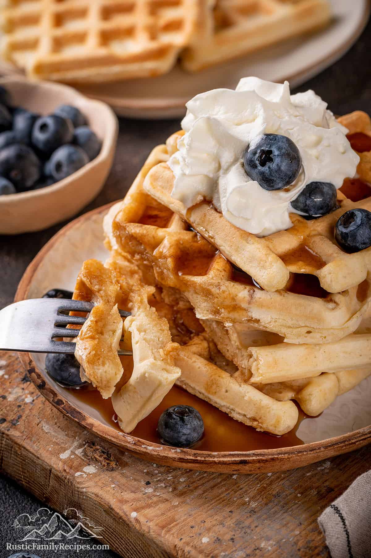 A fork with a bite of sourdough waffles, next to a stack of waffles with whipped cream and berries.