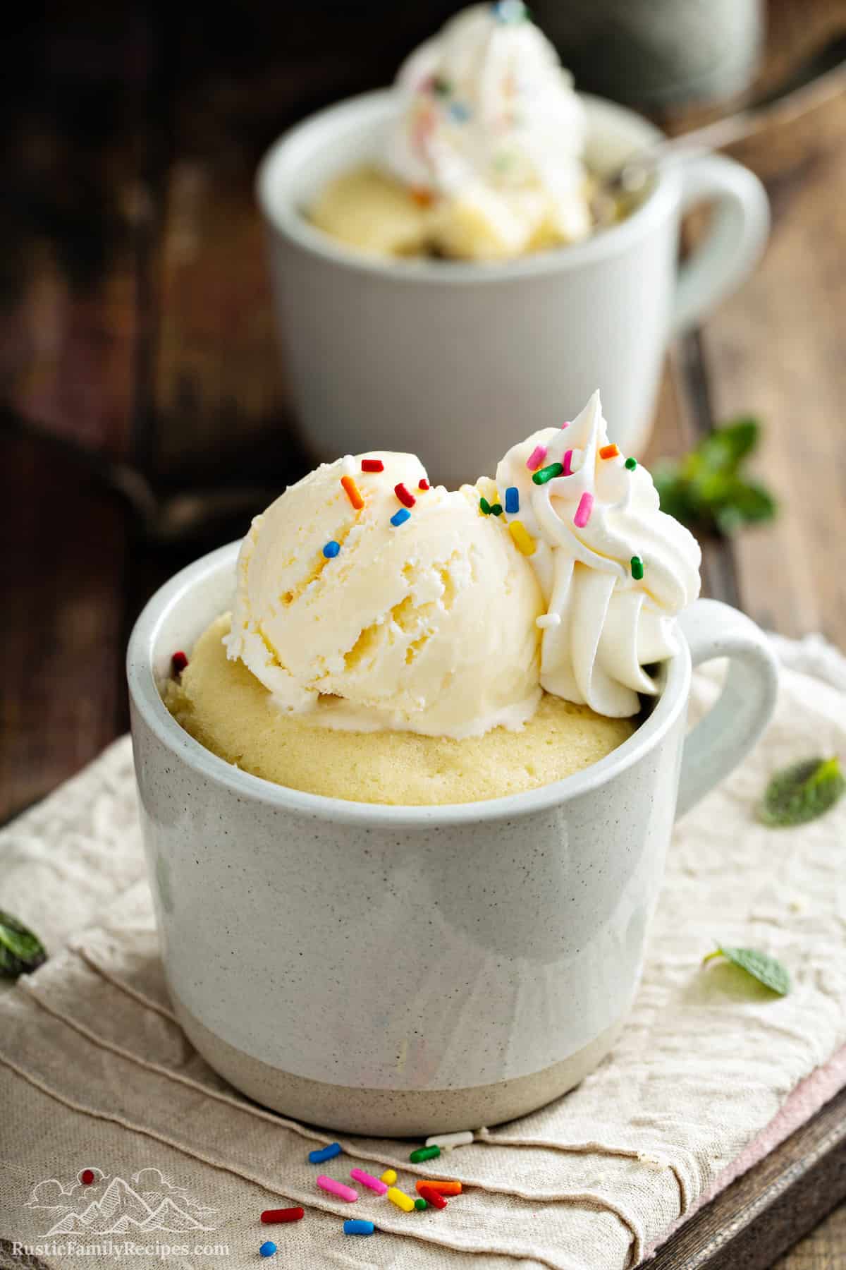 Vanilla mug cake topped with ice cream, whipped cream and sprinkles.