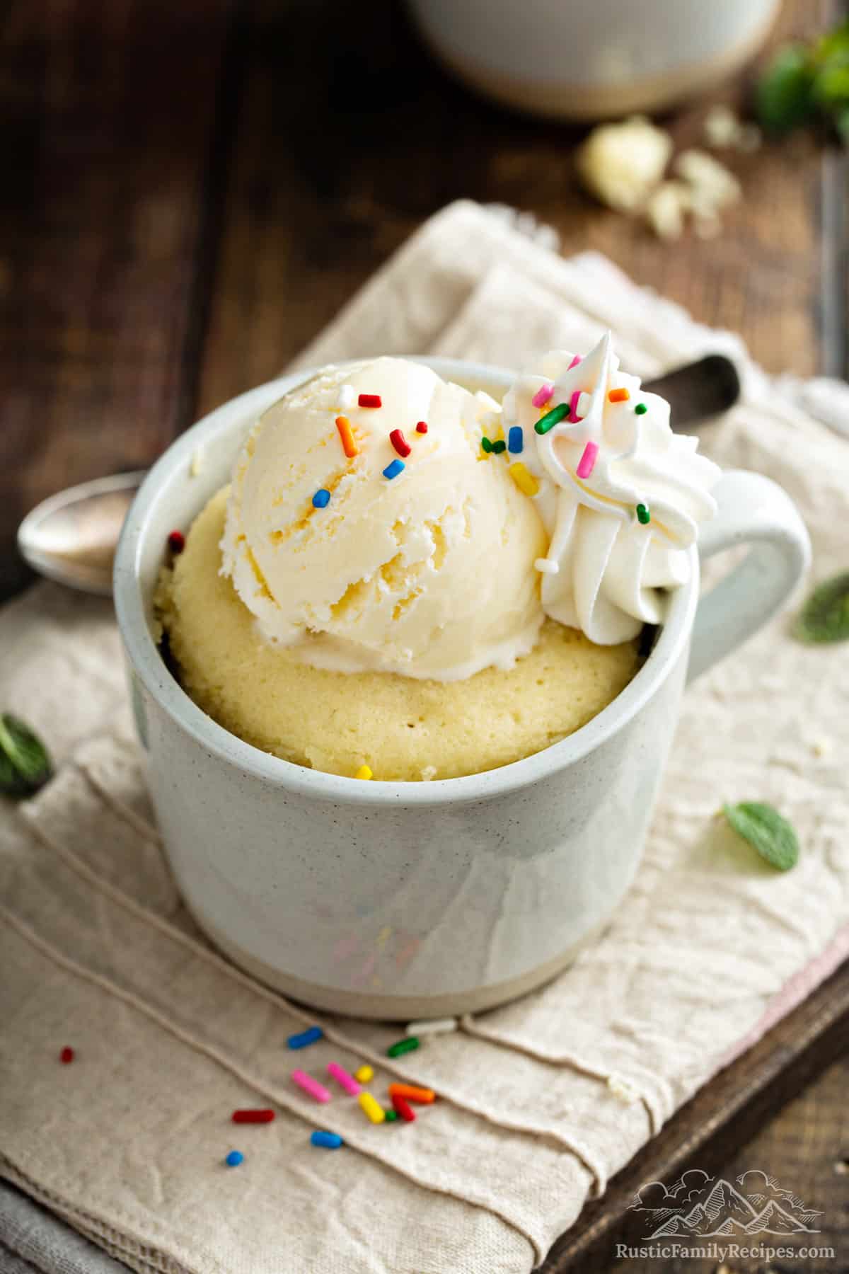 A vanilla cake in a mug topped with ice cream, whipped cream and sprinkles.