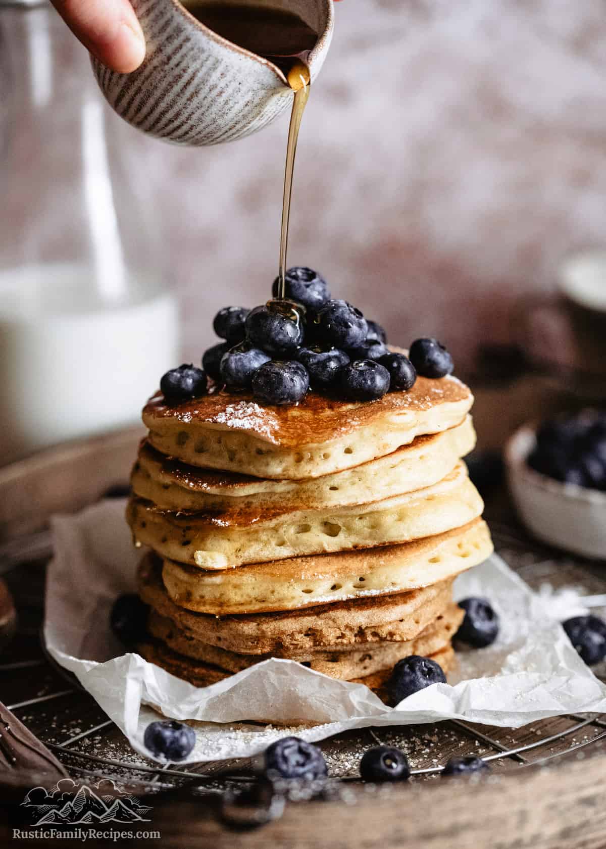 A stack of sourdough pancakes topped with blueberries with syrup being poured on top.