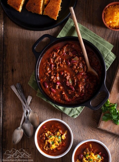 A pot with venison chili next to two bowls with chili and cheese