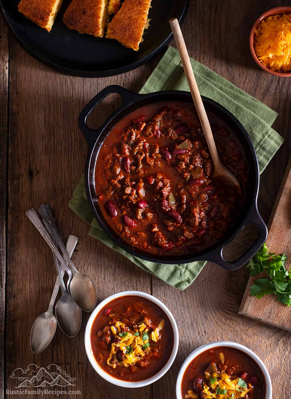 A pot with venison chili next to two bowls with chili and cheese