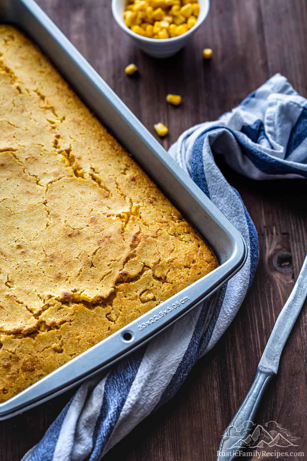 Easy Cheddar Cornbread in a 9 by 13 inch baking pan next to a knife and corn kernels. 