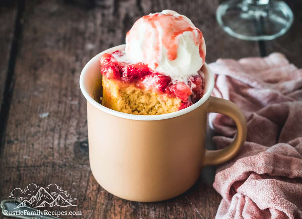 A pink mug on a wood table with vanilla mug cake, strawberries and ice cream