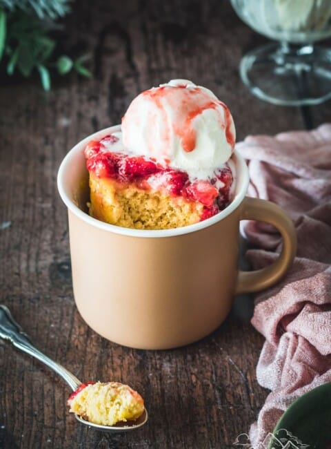 A pink mug on a wood table with vanilla mug cake, strawberries and ice cream