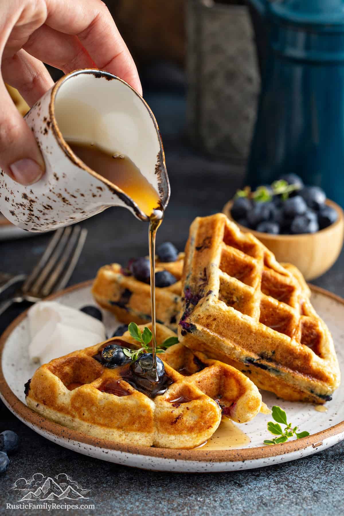 A plate full of fluffy Blueberry Buttermilk Waffles topped with blueberries and being drizzled with maple syrup.