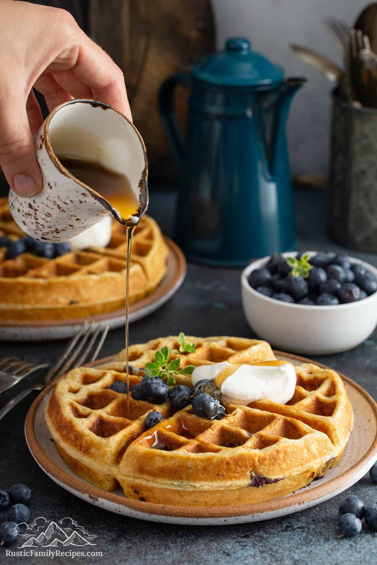 A Blueberry Buttermilk Waffle being topped with blueberries and a drizzle of maple syrup.
