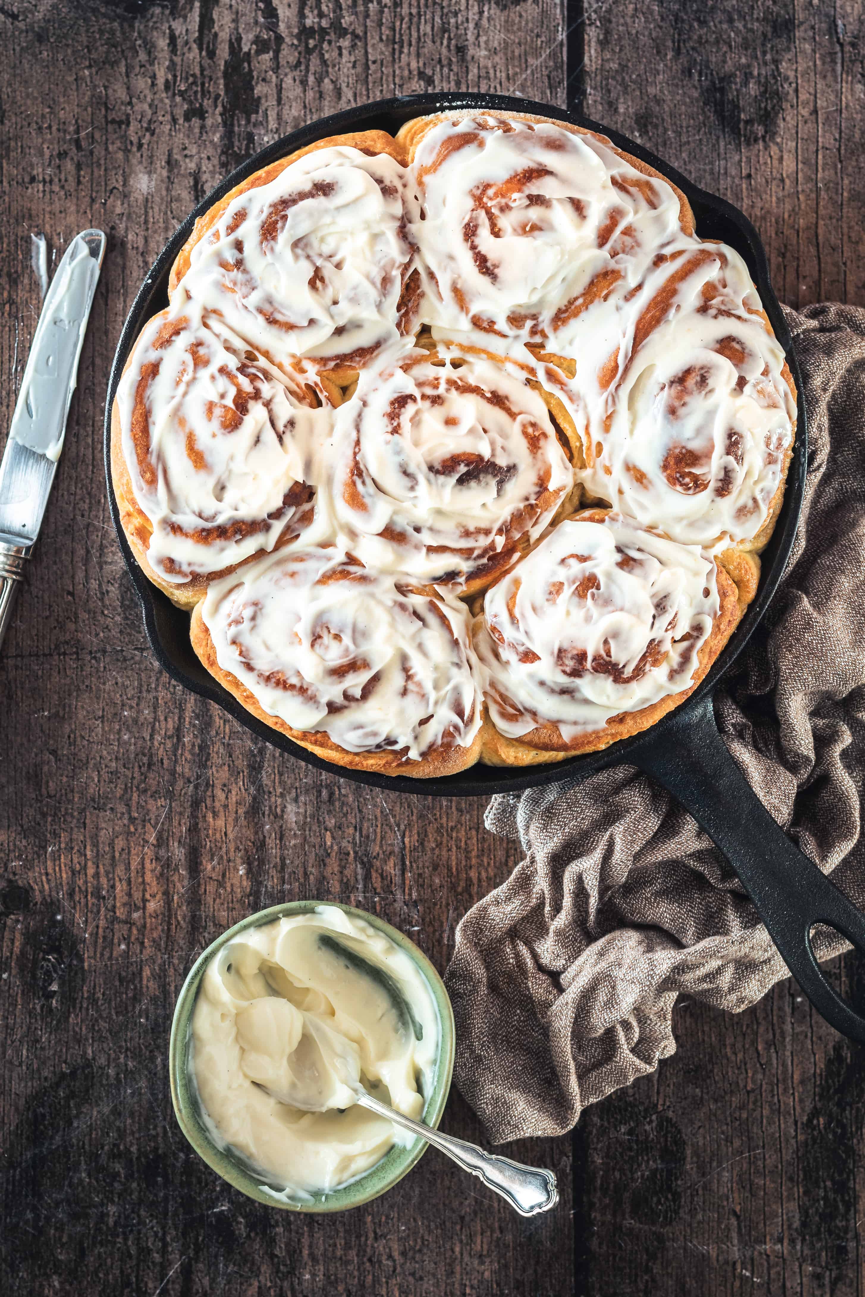 Overhead shot of Sourdough Cinnamon Rolls in skillet with bowl of frosting