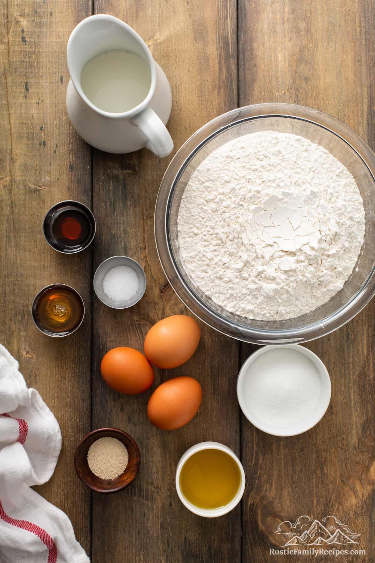 A bowl of flour next to eggs, vanilla, milk and sugar.