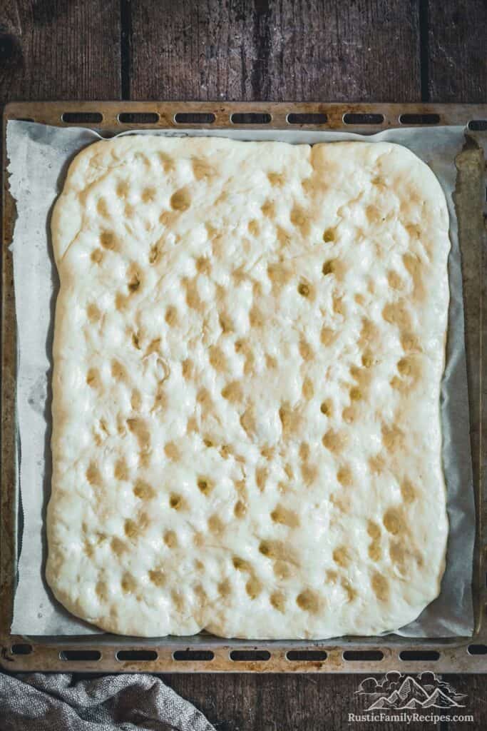 Overhead view of focaccia dough stretched out on baking sheet with finger indentations