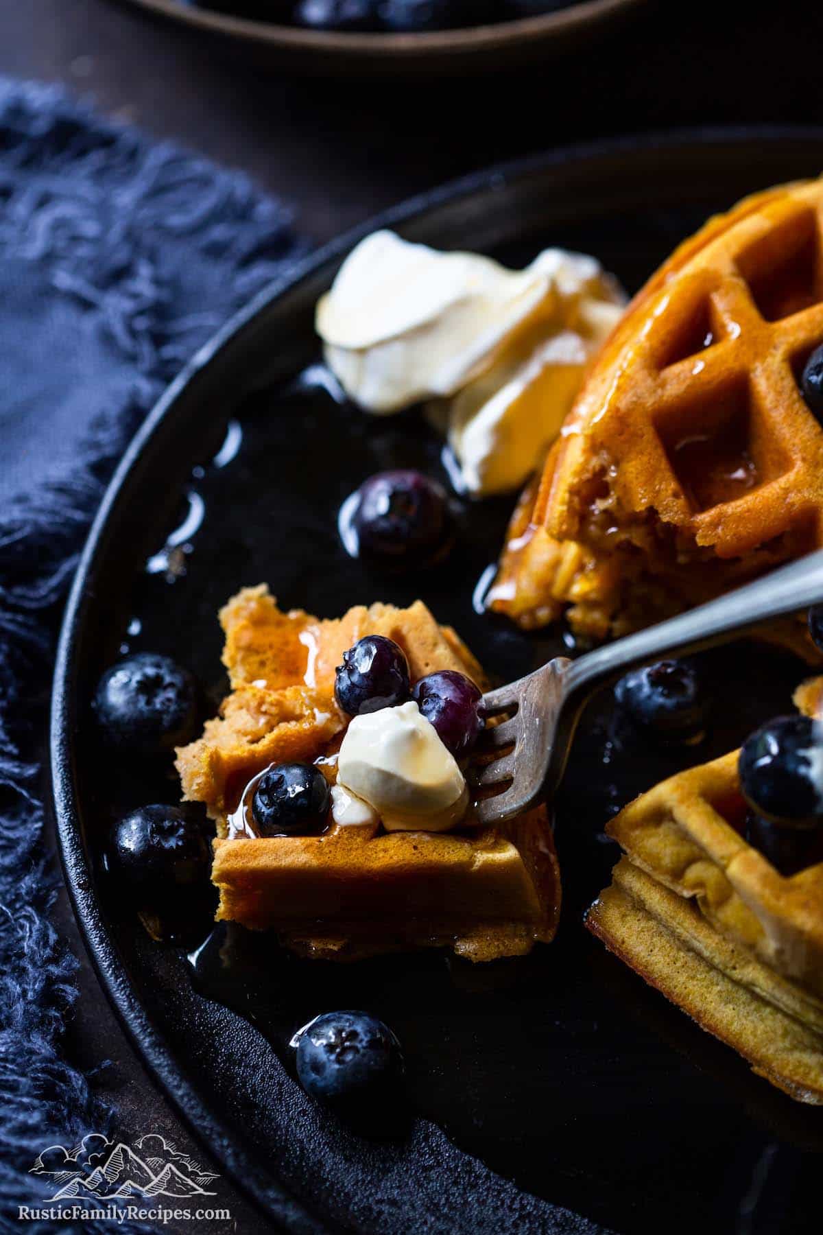 A fork with a bite-full taken out of astack of pumpkin waffles with blueberries, whipped cream and syrup