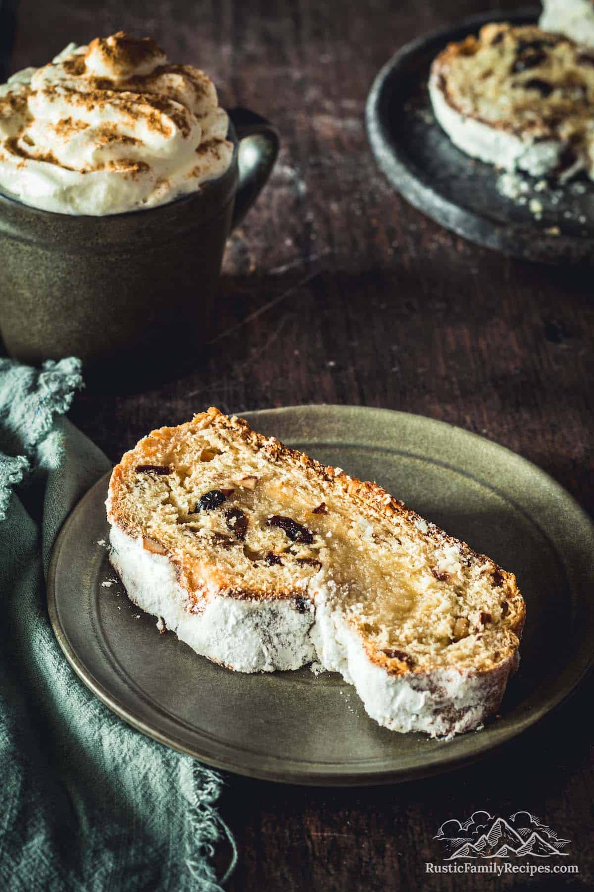 A slice of stollen on a rustic plate with a cup of coffee in the background