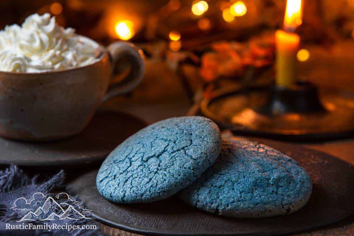 Two blue baby yoda sugar cookies in front of lights with a mug of hot chocolate in the background