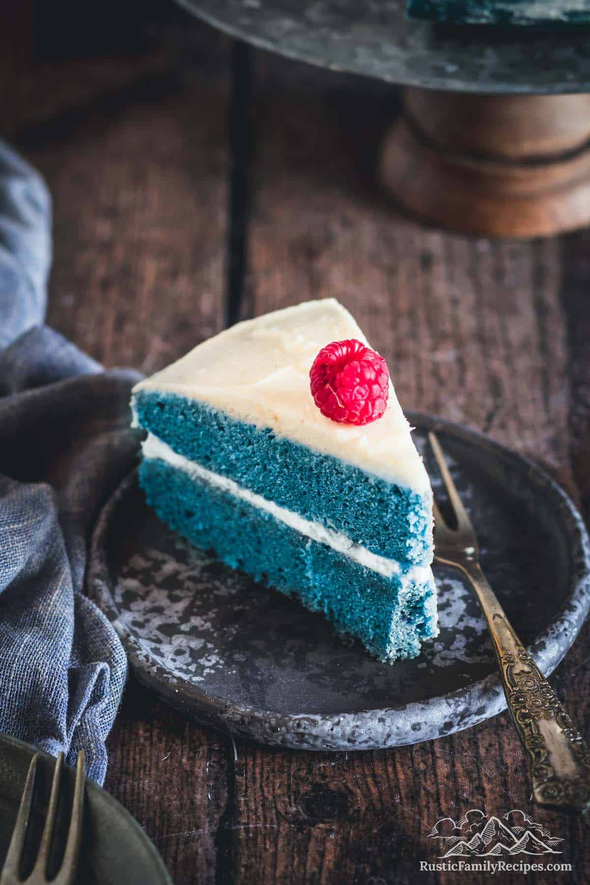 A sliced of blue cake frosted with vanilla frosting and topped with a raspberry