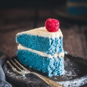 A slice of blue cake with a bite taken out, a raspberry on top of the slice