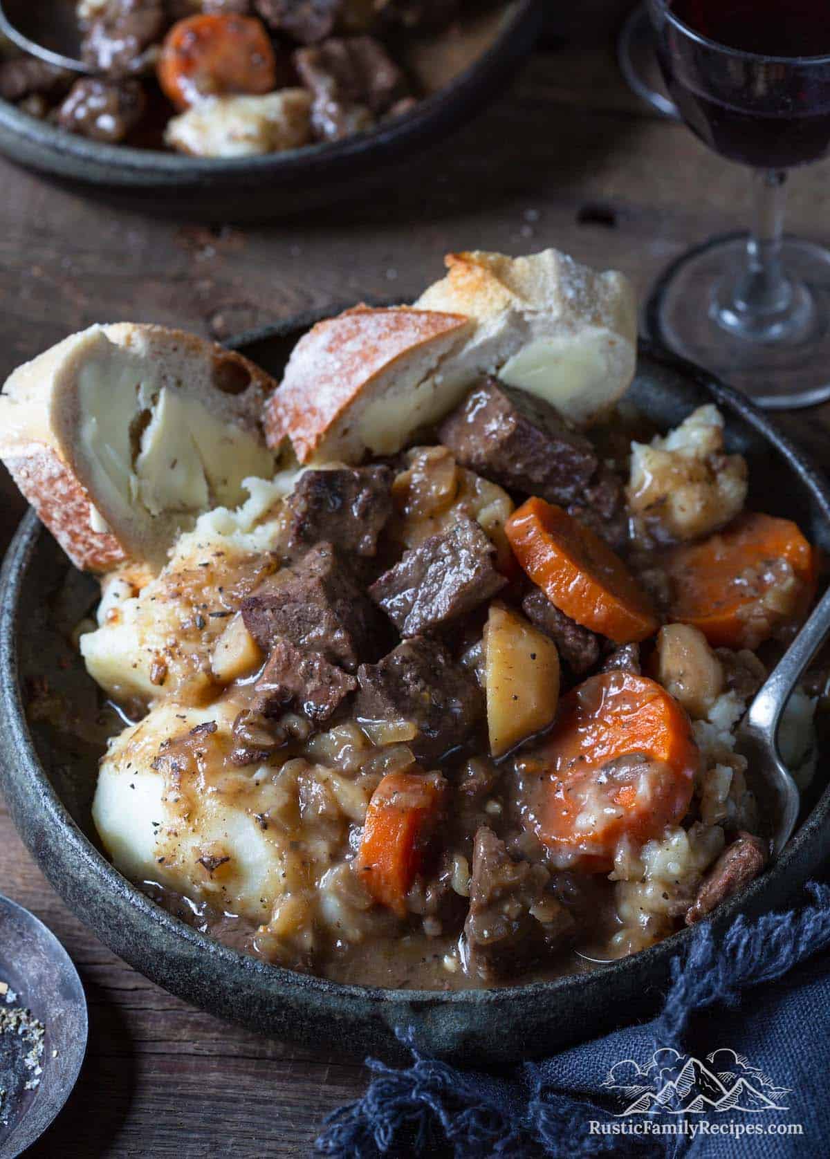 A bowl filled with venison stew, mashed potatoes and crusty bread slices