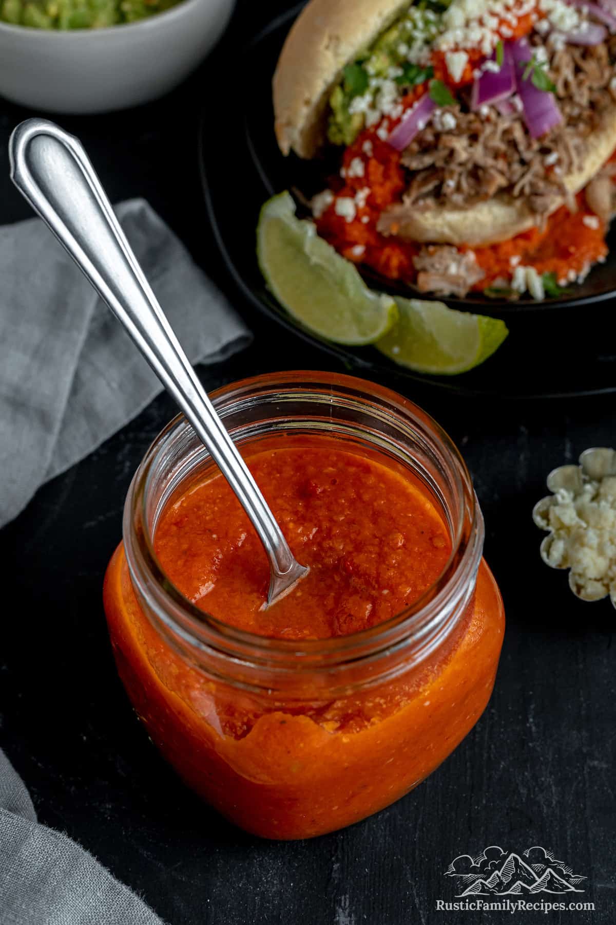 A mason jar filled with red chili sauce with a spoon, next to a torta sandwich