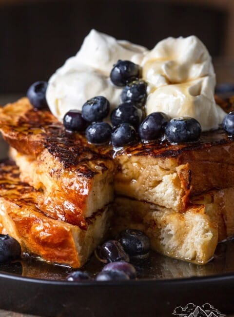 Two slices of challah french toast with a slice taken out, whipped cream and blueberries on top