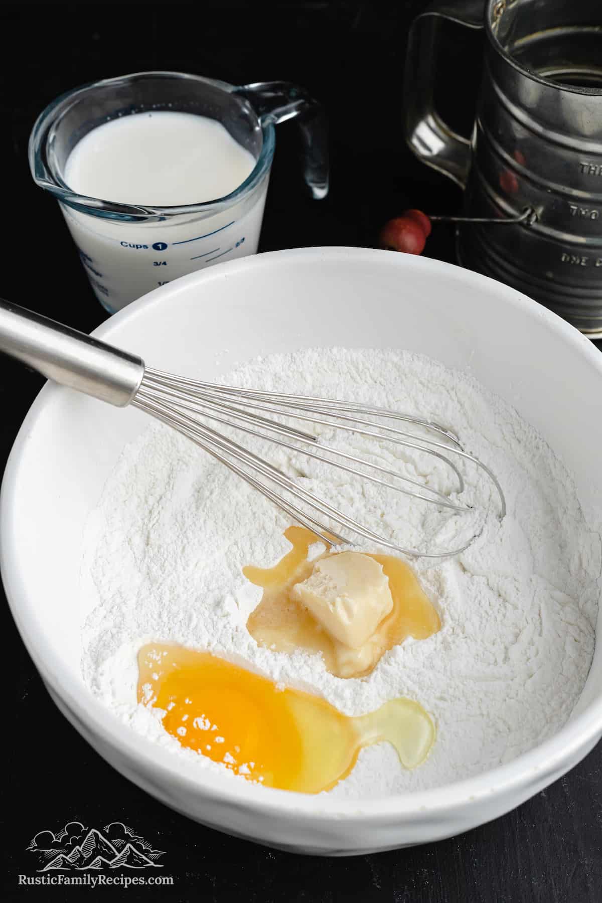 A white bowl with flour, butter, eggs and a whisk