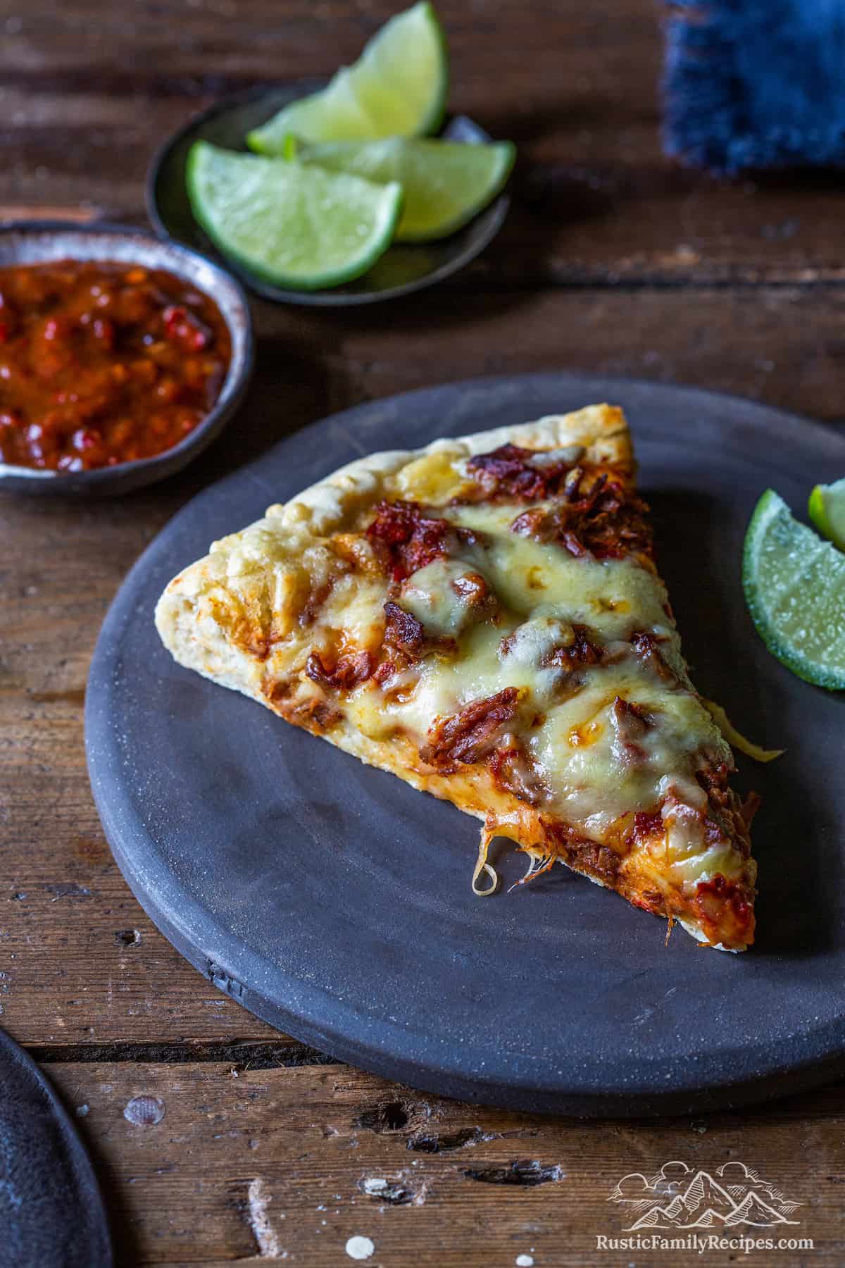 A slice of birria pizza on a plate with lime wedges