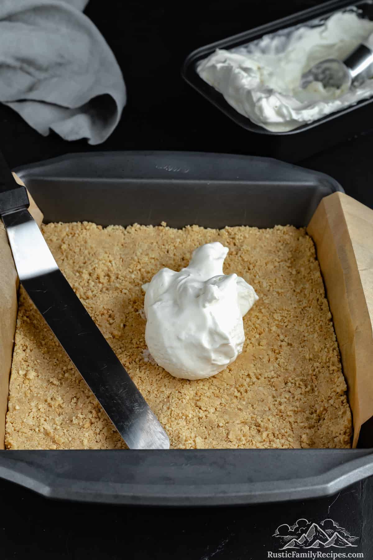 A scoop of vanilla ice cream is ready to be spread over the Golden Oreo crust that's been pressed into the bottom of a lined baking pan.