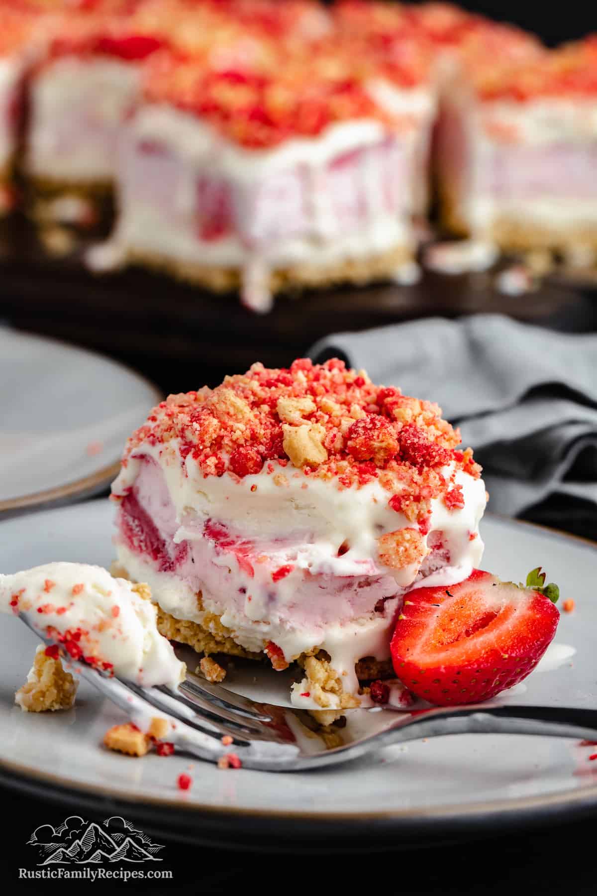 A fork cuts into a strawberry shortcake ice cream bar on a plate next to half of a fresh strawberry, with more bars in the background.