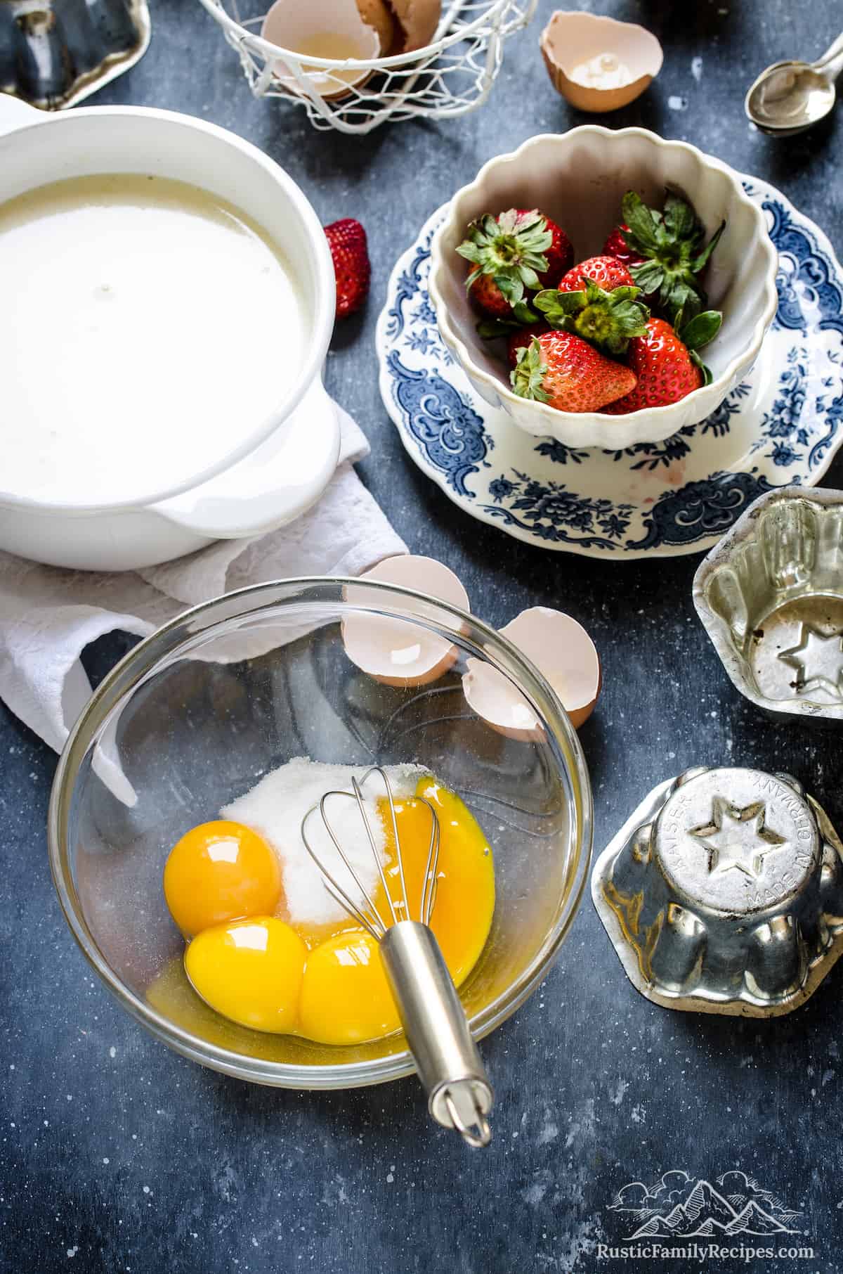 Egg yolks are whisked together with sugar in a glass mixing bowl, next to the pudding cream mixture and a bowl of strawberries.