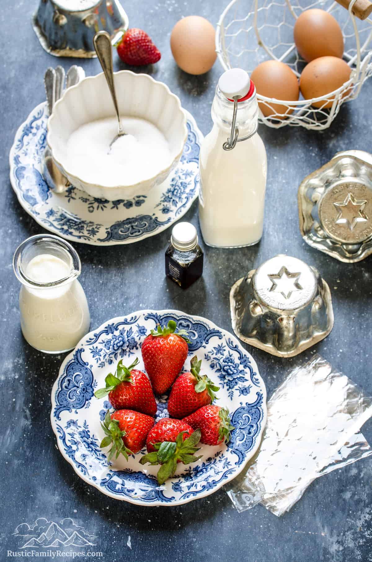 A bowl of strawberries next to a bowl of vanilla cream mixture and a bowl of eggs, next to scattered mini bundt cake tins.