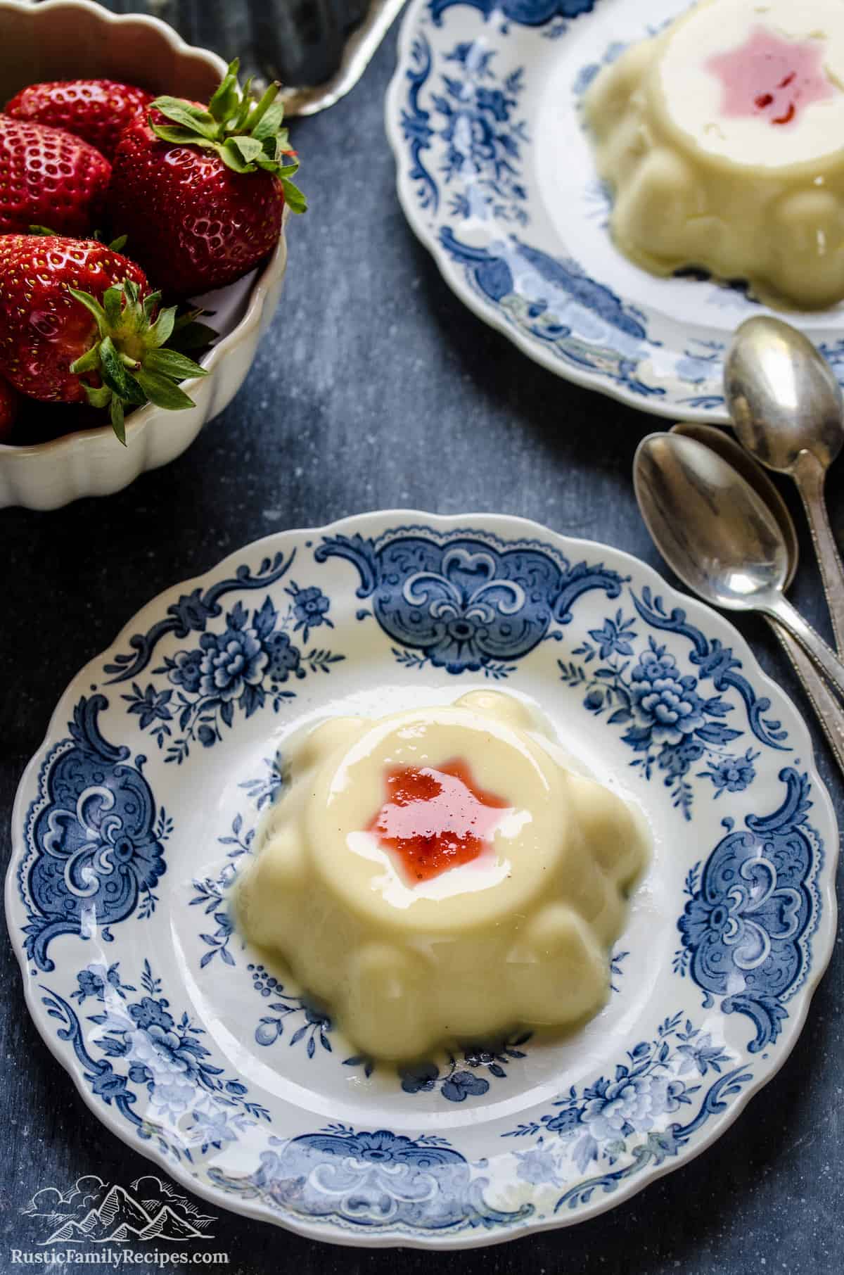 Top view of two vanilla budinos on Delftware plates, next to a bowl of strawberries.