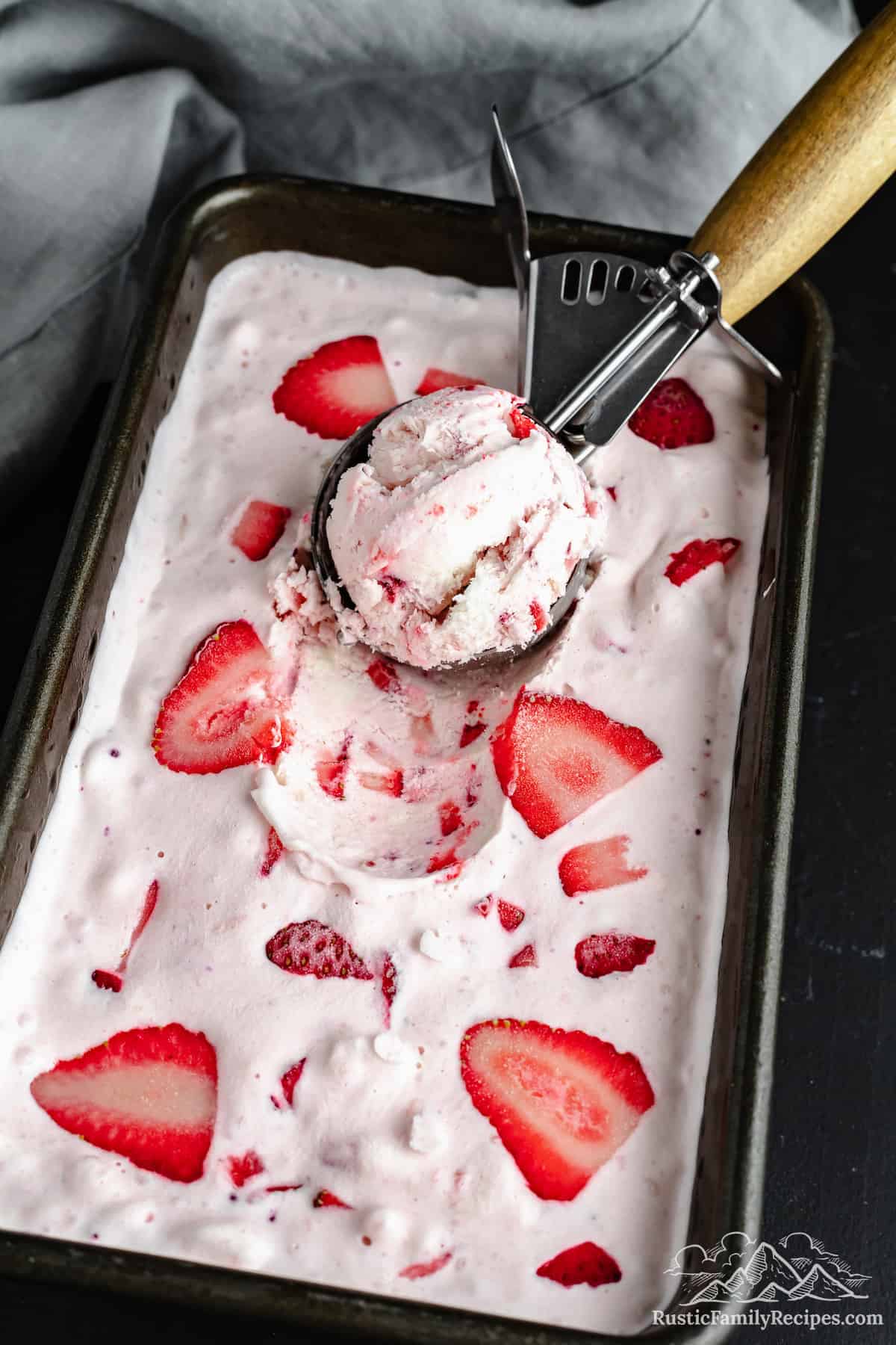An ice cream scoop picks up a ball of ice cream from a pan full of homemade strawberry ice cream.