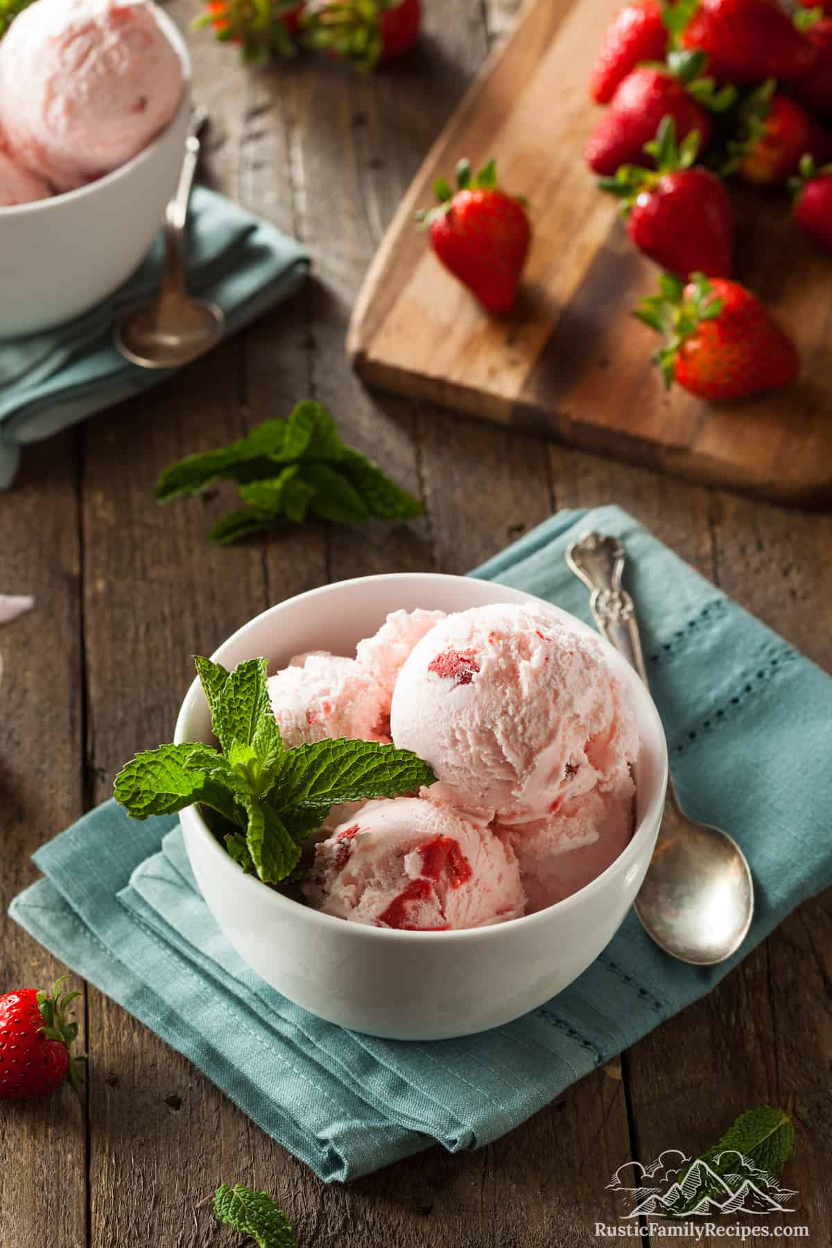 Scoops of strawberry ice cream in a bowl garnished with mint, with strawberries scattered on a wooden board in the background.
