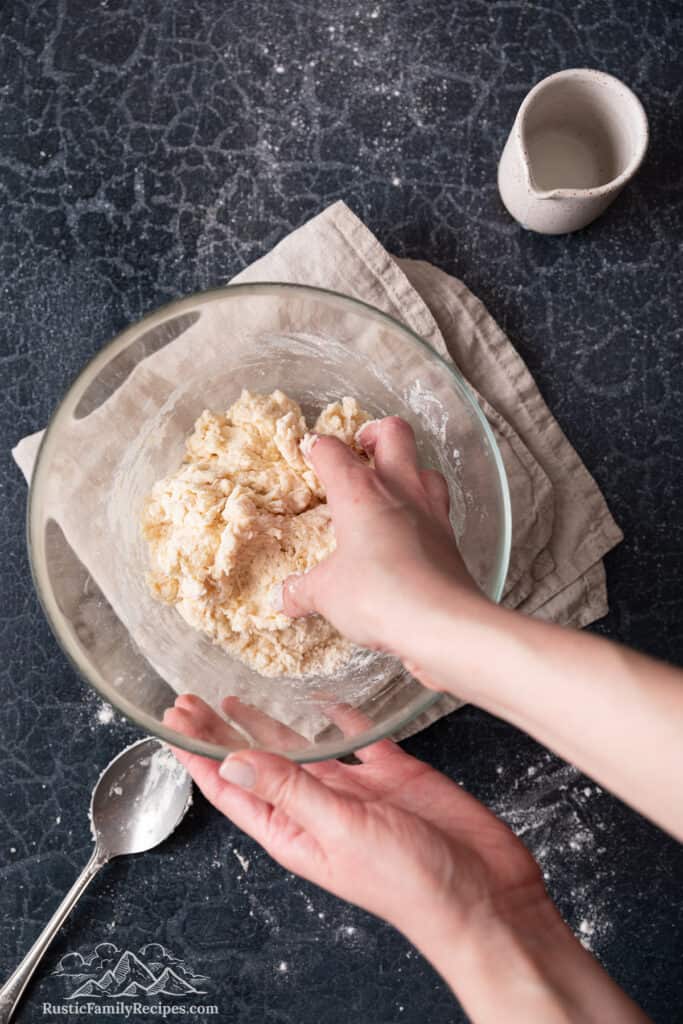 A hand kneads together the ingredients for biscuit dough in a mixing bowl.
