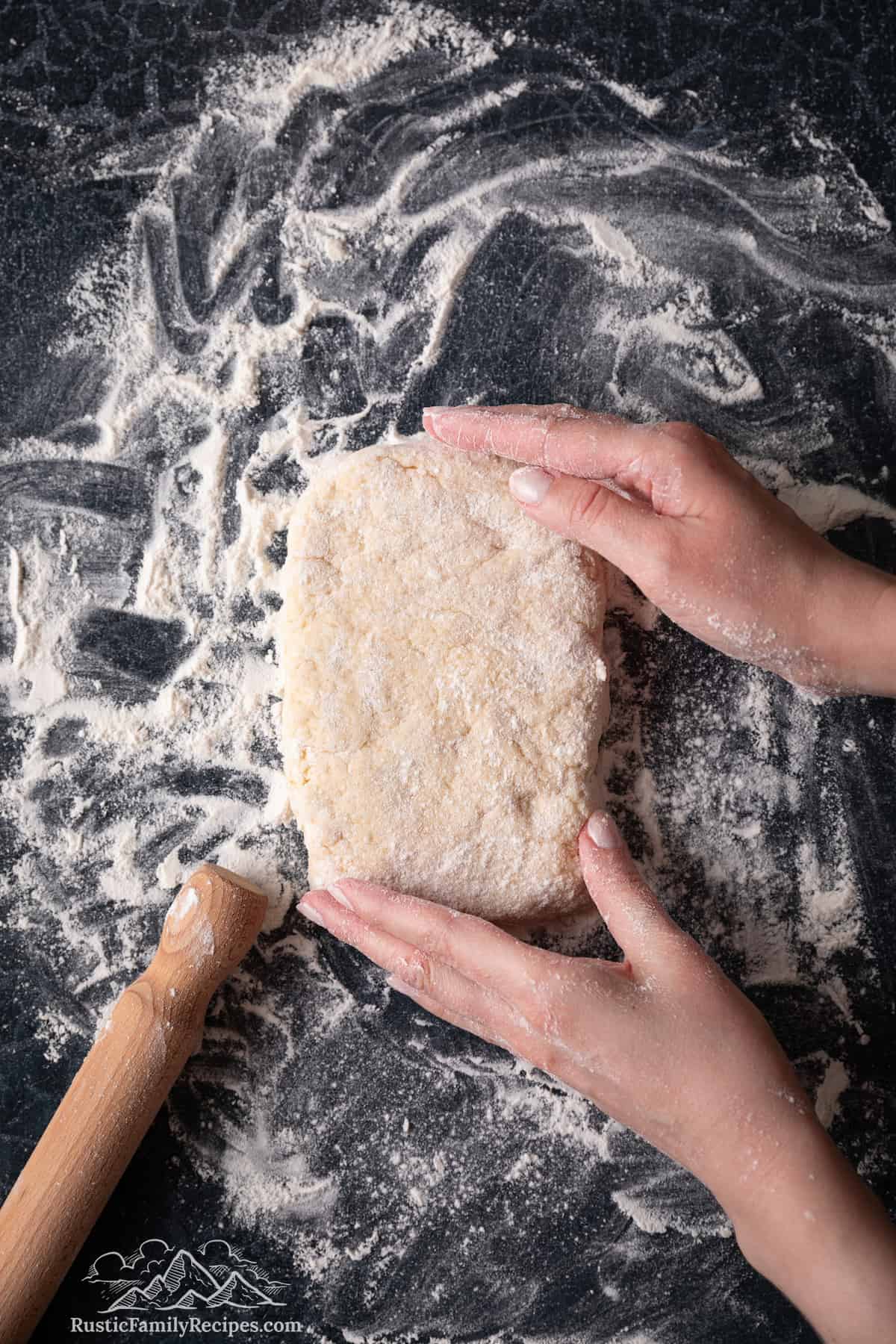 Two hands shape shortcake biscuit dough into a rectangle on a floured surface.