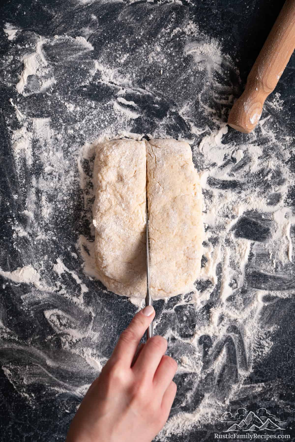 A hand uses a knife to cut a rectangle of shortcake biscuit dough in half lengthwise.