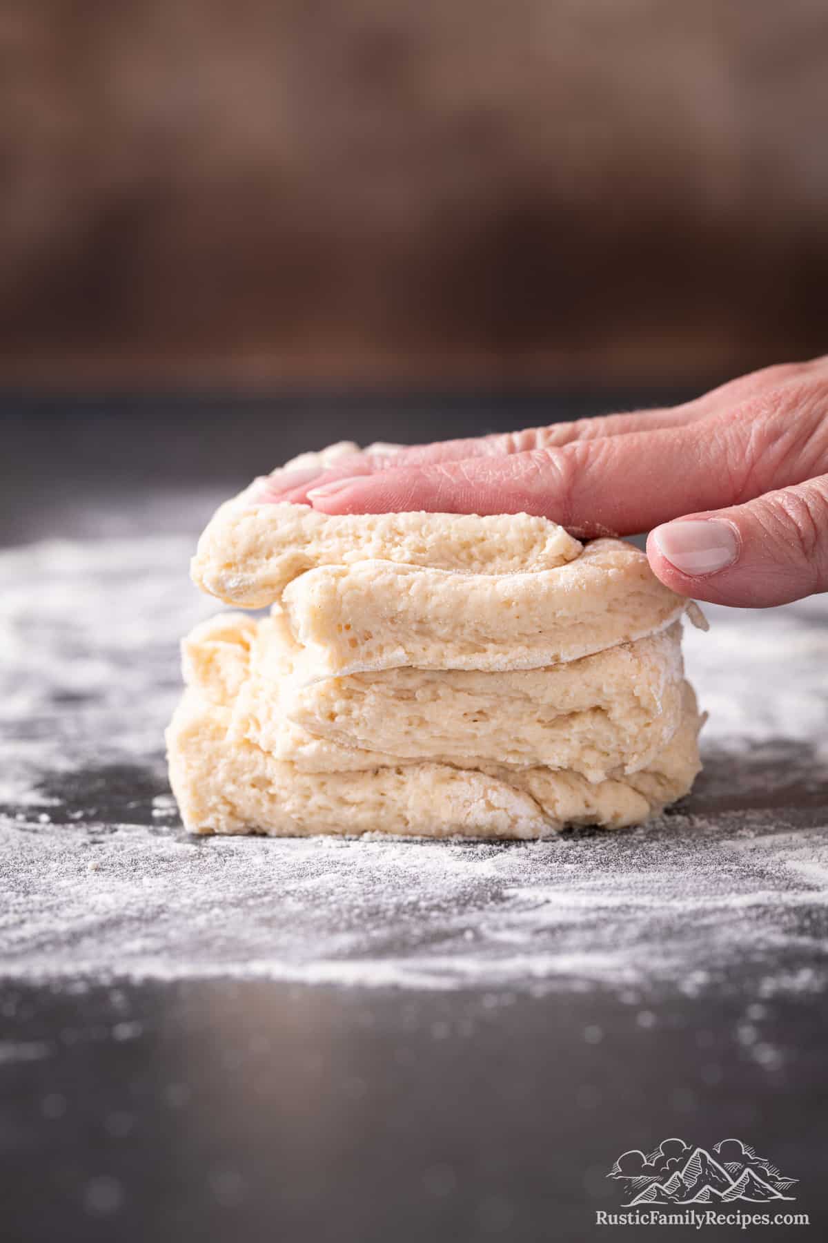 A hand presses a stack of shortcake dough pieces down onto a floured surface.