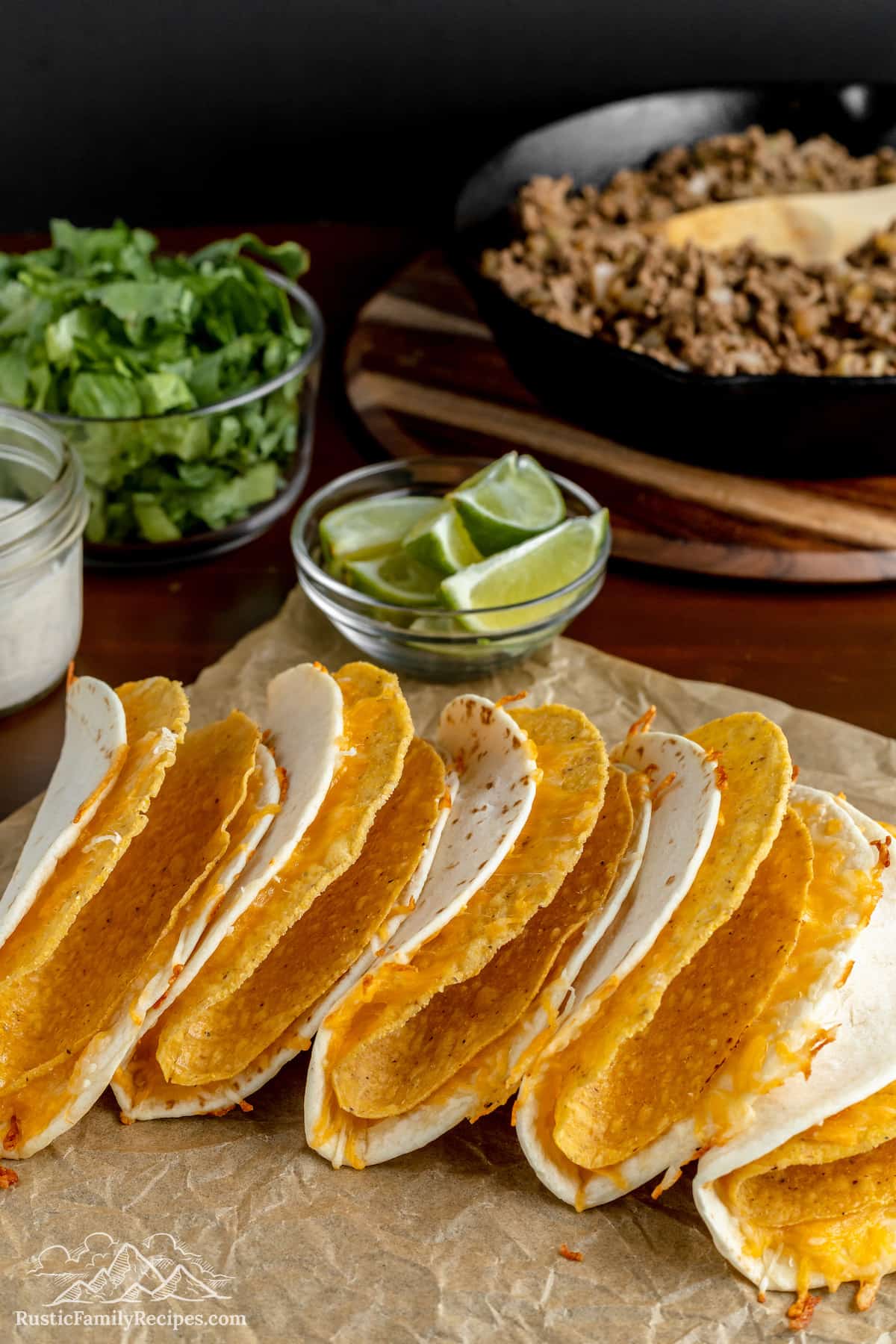 Double-layered taco shells in a row with a skillet of taco meat and bowls with limes and lettuce in the background.