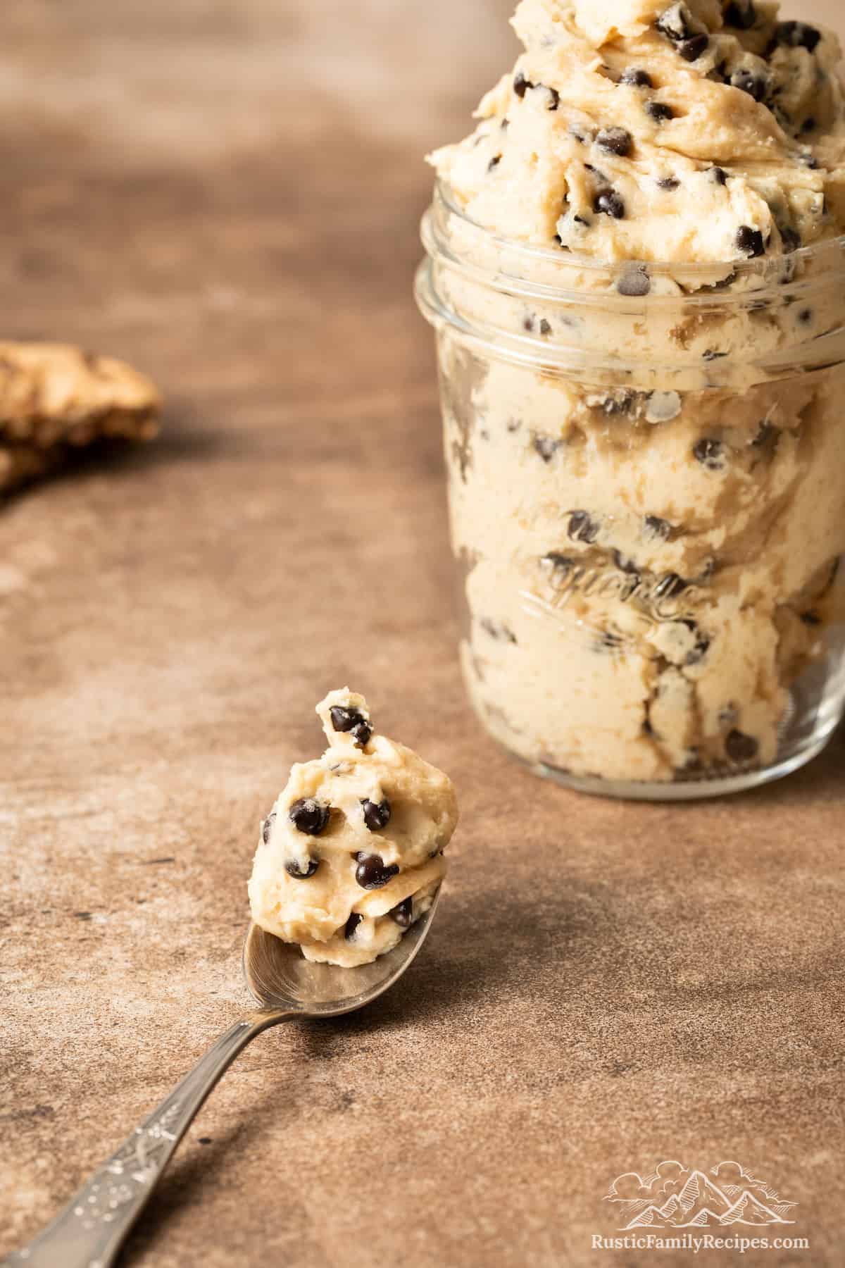A mason jar with edible chocolate chip cookie dough and spoonful bedside it