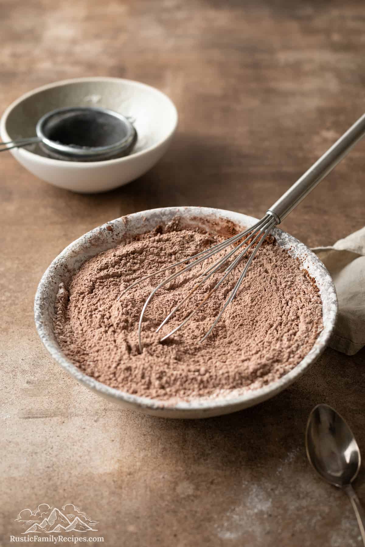Flour, cocoa powder, and dry ingredients whisked together in a large bowl, with a sift in a small bowl in the background.