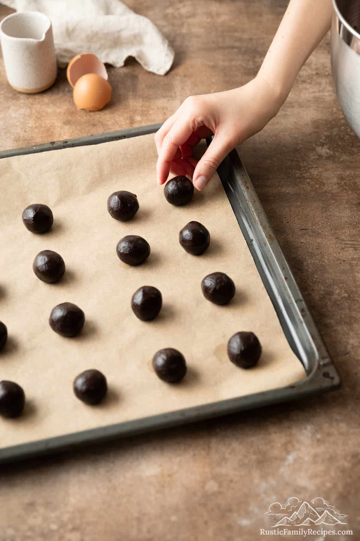 A hand places dark chocolate cookie dough balls out in rows on a parchment-lined baking sheet.