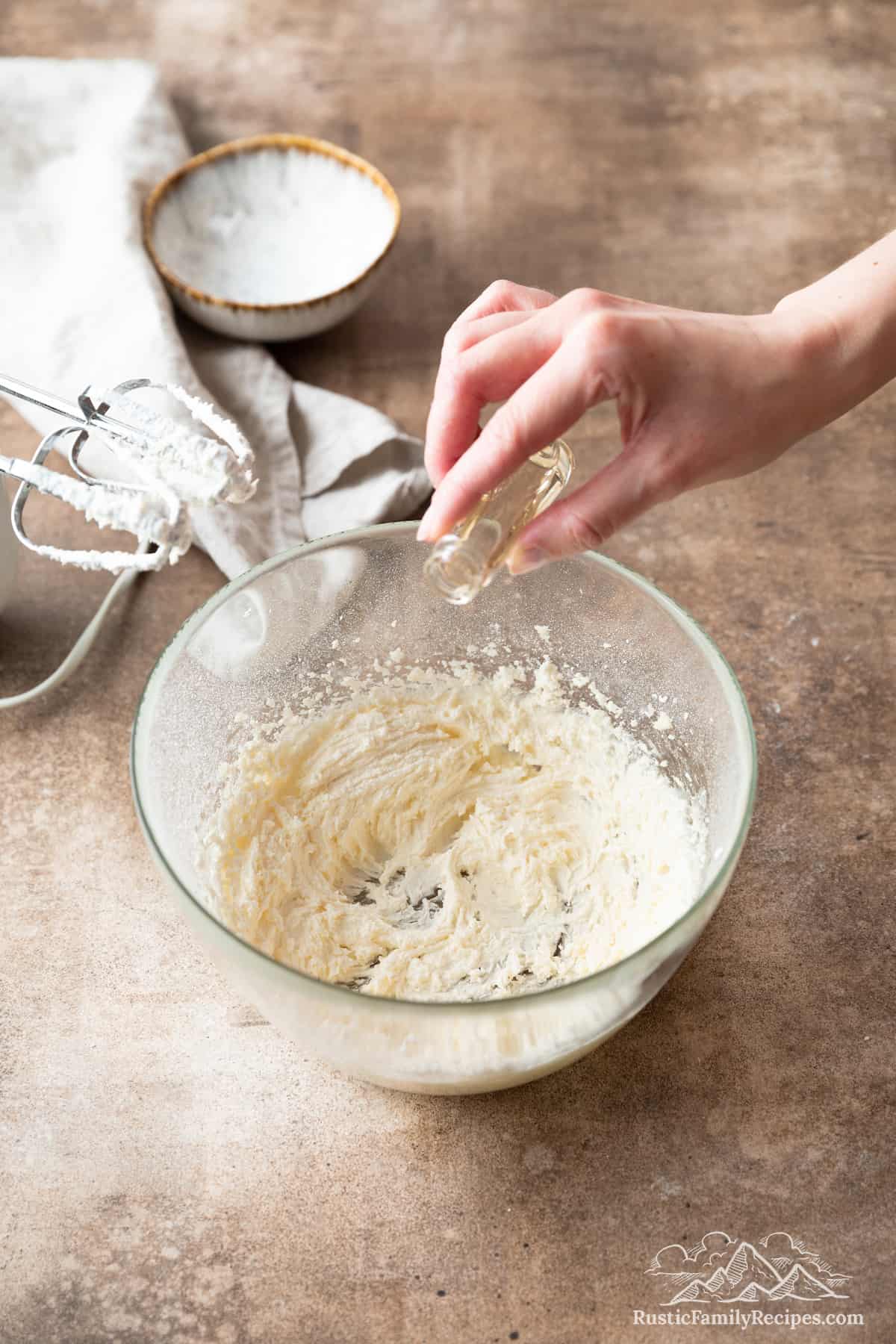 A hand pours vanilla extract from a small bottle into a bowl of powdered sugar creamed with butter, next to a hand mixer.